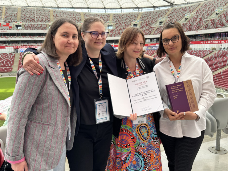 Cztery pracownice działu wydawniczego stolą na stadionie narodowym w Warszawie. Osoba w środku trzyma nagrodę ACADEMIA w formie papierowego dyplomu A4, osoba obok trzyma w ręku nagrodzoną książkę.  