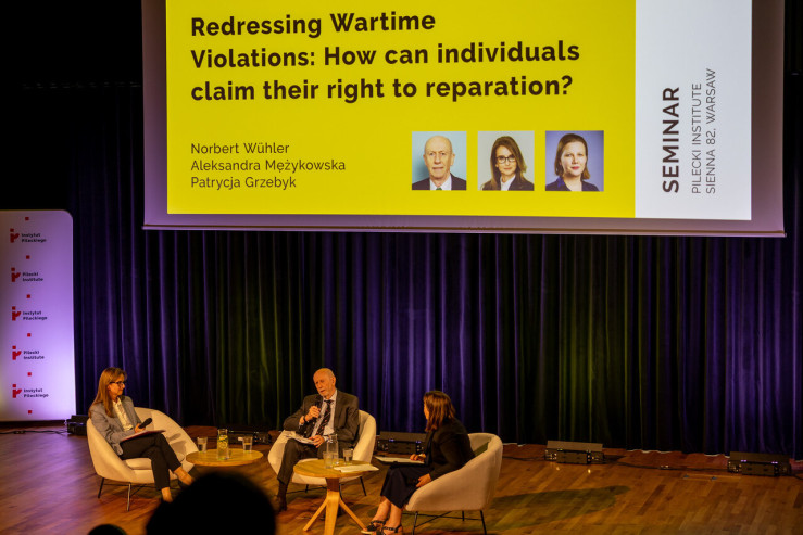 Wide shot of the stage at the Pilecki Institute showing three panelists seated in white armchairs below a large presentation slide titled "Redressing Wartime Violations: How can individuals claim their right to reparation?".