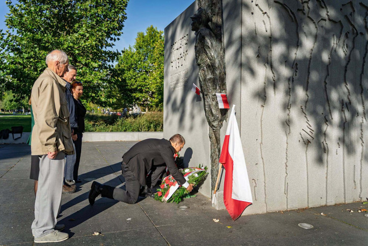 Dyrektor Madaj w ciemnym garniturze klęczy i składa wieniec pod pomnikiem rotmistrza Witolda Pileckiego w obecności stojącej obok grupy osób.