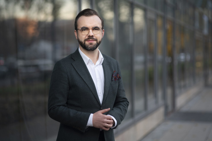 Portret mężczyzny (Michał Kosiorek) w okularach, ciemnozielonej marynarce i białej koszuli, stojącego przed przeszklonym budynkiem.