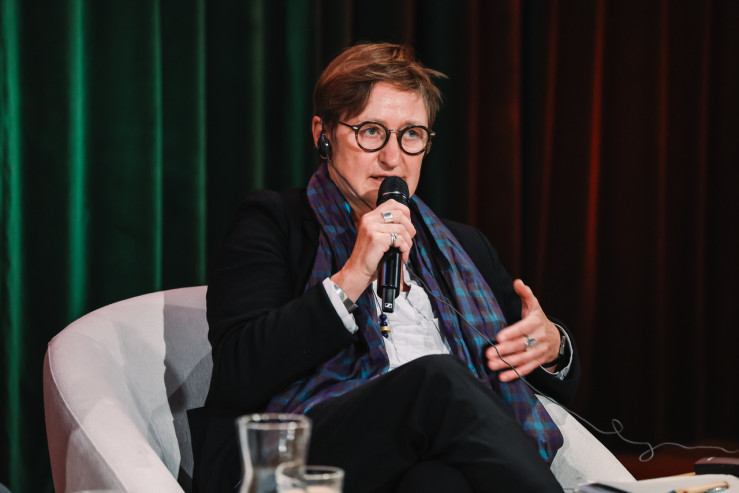 Kobieta w okularach, marynarce i szalu przemawia do mikrofonu podczas panelu dyskusyjnego.