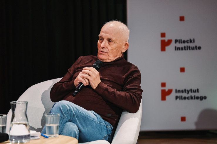 Mężczyzna w brązowym swetrze wypowiada się do mikrofonu na tle ścianki Instytutu Pileckiego.
