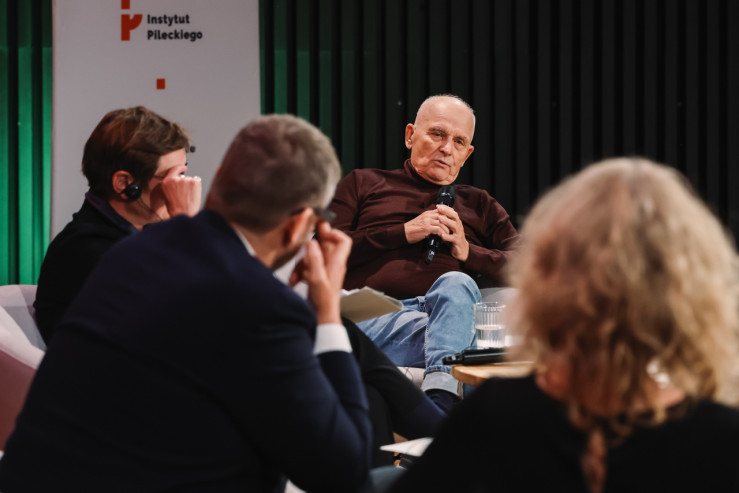 Ujęcie zza pleców słuchaczy i innych uczestników panelu w stronę mężczyzny siedzącego w centrum, który mówi do mikrofonu. W tle widoczna jest ścianka prasowa z logo Instytutu Pileckiego.
