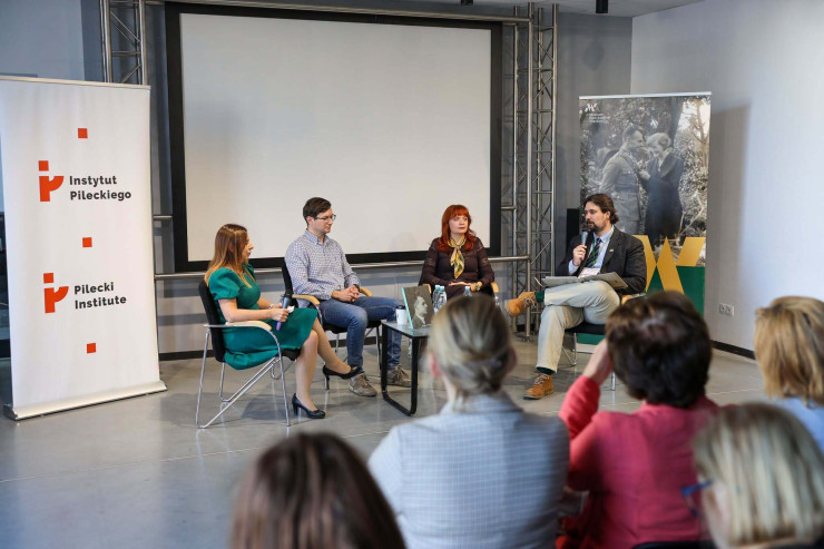 Panel dyskusyjny z udziałem czterech osób siedzących w fotelach na scenie przed publicznością. Po lewej stronie stoi roll-up Instytutu Pileckiego, a po prawej banner Muzeum Dom Rodziny Pileckich.