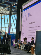 scena konferencyjna na której siedzi w fotelach 5 panelistów, po lewej stronie przy pulpicie do mikrofonu mówi kobieta, z tyłu wielki ekran ok 4 na 7 metrów.