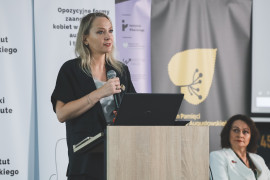 Prelegentka konferencji naukowej przemawia do mikrofonu przy mównicy z laptopem. W tle widoczne są banery informacyjne z tytułem konferencji „Opozycyjne formy zaangażowania kobiet w systemach autorytarnych i totalitarnych XX wieku” oraz logotypy Instytutu Pileckiego i programu Ministerstwa Nauki „Wektory Nauki”.