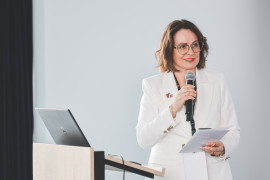 Prelegentka przemawiająca podczas międzynarodowej konferencji naukowej. Prelegentka w okularach i białym żakiecie stoi przy mównicy z laptopem, trzymając w dłoniach mikrofon oraz program konferencji.