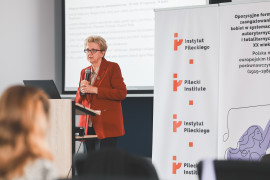 Prelegentka w okularach i rdzawym żakiecie wygłasza referat podczas konferencji. Stoi przy mównicy, trzymając mikrofon w obu dłoniach. W tle widoczne banery z logotypem Instytutu Pileckiego i tytułem wydarzenia oraz ekran z fragmentem prezentacji naukowej.
