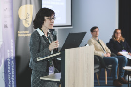 Prelegentka w okularach, szarym żakiecie i kraciastej spódnicy wygłasza referat przy mównicy z laptopem. W tle widoczne rollupy z logotypami programu „Wektory Nauki” oraz Domu Pamięci Obławy Augustowskiej – filii Instytutu Pileckiego. Na drugim planie siedzą pozostali członkowie panelu tematycznego, oczekujący na swoje wystąpienia.