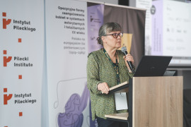 Prelegentka o siwych włosach z prostą grzywką, w okularach i zielonej, wzorzystej bluzce, wygłasza referat przy mównicy z laptopem. W tle widoczne rollupy z logotypami programu „Wektory Nauki” oraz Domu Pamięci Obławy Augustowskiej – filii Instytutu Pileckiego. Na banerach widnieje tytuł międzynarodowej konferencji naukowej o historii opozycyjnych form zaangażowania kobiet w XX wieku.