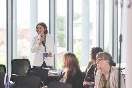 Uczestniczka dyskusji w białym żakiecie, stojąc wśród publiczności zadaje pytanie przez mikrofon. Na pierwszym planie widoczne są siedzące słuchaczki konferencji. W tle znajdują się wysokie, przeszklone ściany, po których spływają krople deszczu.