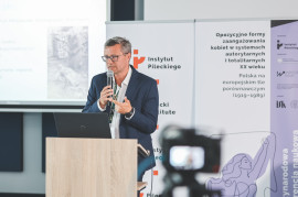 Prelegent w okularach, białej koszuli i granatowej marynarce wygłasza referat przy mównicy z laptopem marki Dell. Mężczyzna trzyma mikrofon w prawej dłoni, a lewą gestykuluje; na lewym nadgarstku ma czarny zegarek. W tle widoczny jest ekran z prezentacją naukową przedstawiającą archiwalne, czarno-białe zdjęcie a także rollupy z pełnym tytułem konferencji i logotypami Instytutu Pileckiego, programu „Wektory Nauki”, Ministra Nauki i Szkolnictwa Wyższego oraz Instytutu Studiów Kobiecych. Pierwszy plan jest nieostry i przedstawia kamerę rejestrującą wystąpienie.