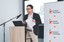 Prelegent o krótkich ciemnych włosach, w okularach, jasnej koszuli w delikatną kratkę i ciemnej marynarce, wygłasza referat przy drewnianej mównicy. W dłoniach trzyma wydrukowany tekst wystąpienia oraz butelkę wody. Przed nim stoi mikrofon na statywie oraz laptop marki Dell. W tle widoczny pionowy rollup Instytutu Pileckiego oraz ekran z wyświetlonym archiwalnym, czarno-białym zdjęciem.