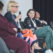 Uczestnicy panelu naukowego podczas dyskusji. Po lewej stronie, częściowo w kadrze, widoczna postać w purpurowym komplecie. Obok niej prelegentka o krótkich blond włosach i w okularach mówi do mikrofonu. Dalej siedzi uczestniczka w ciemnej marynarce we wzory kwiatów i liści, z barwną tkaniną w geometryczne wzory na kolanach. Po prawej prelegent w okularach i błękitnym krawacie w skośne paski. Wszyscy paneliści mają na szyi zielone smycze z identyfikatorami.