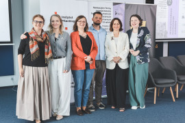 Zdjęcie grupowe sześciorga organizatorów i prelegentów na tle banerów konferencyjnych. Od lewej stoją: prelegentka w kolorowym szalu i długiej beżowej spódnicy, uczestniczka w szarym kardiganie i białych spodniach, osoba w koralowej marynarce i jeansach, mężczyzna w błękitnej koszuli, kobieta w białym żakiecie oraz prelegentka w ciemnej marynarce we wzory kwiatów i liści. W tle widoczne rollupy z logotypami programu „Wektory Nauki”, Instytutu Pileckiego oraz Instytutu Studiów Kobiecych.