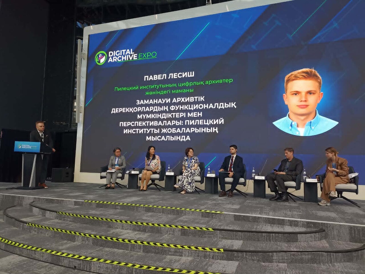 scena konferencyjna na której siedzi w fotelach 5 panelistów, po lewej stronie przy pulpicie do mikrofonu mówi kobieta, z tyłu wielki ekran ok  4 na 7 metrów. 