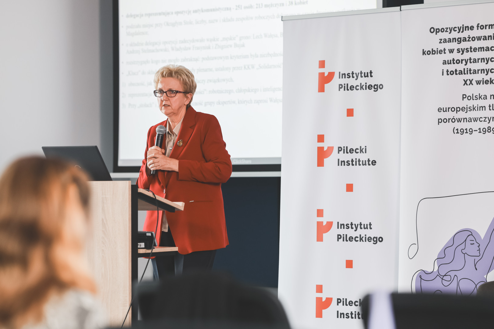 Prelegentka w okularach i rdzawym żakiecie wygłasza referat podczas konferencji. Stoi przy mównicy, trzymając mikrofon w obu dłoniach. W tle widoczne banery z logotypem Instytutu Pileckiego i tytułem wydarzenia oraz ekran z fragmentem prezentacji naukowej.