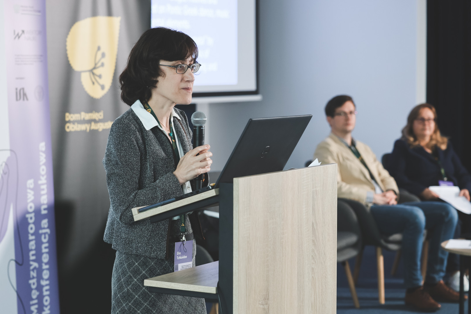 Prelegentka w okularach, szarym żakiecie i kraciastej spódnicy wygłasza referat przy mównicy z laptopem. W tle widoczne rollupy z logotypami programu „Wektory Nauki” oraz Domu Pamięci Obławy Augustowskiej – filii Instytutu Pileckiego. Na drugim planie siedzą pozostali członkowie panelu tematycznego, oczekujący na swoje wystąpienia.