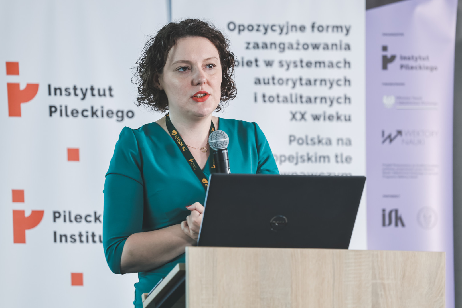 Prelegentka z ciemnymi, kręconymi włosami, w morskiej sukience, wygłasza referat przy mównicy z laptopem. W tle widoczne rollupy z tytułem międzynarodowej konferencji o zaangażowaniu kobiet w XX wieku oraz logotypami Instytutu Pileckiego i programu „Wektory Nauki”.