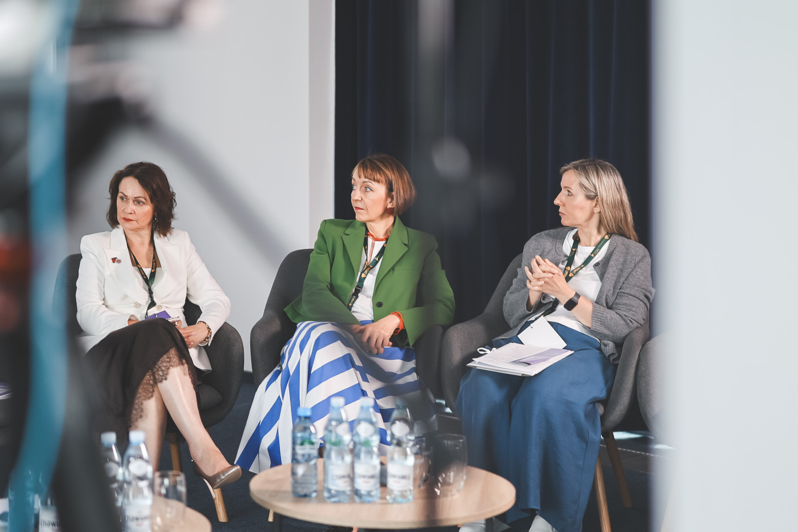 Uczestniczki panelu tematycznego siedzące w fotelach podczas słuchania wystąpienia. Od lewej: prelegentka w białej marynarce i czarnej spódnicy, prelegentka w zielonym żakiecie i spódnicy w biało-niebieskie pasy oraz prelegentka w szarym kardiganie. Na stoliku przed nimi butelki wody i szklanki. Widok zza rozmazanego sprzętu technicznego na pierwszym planie. Konferencja programu „Wektory Nauki”.