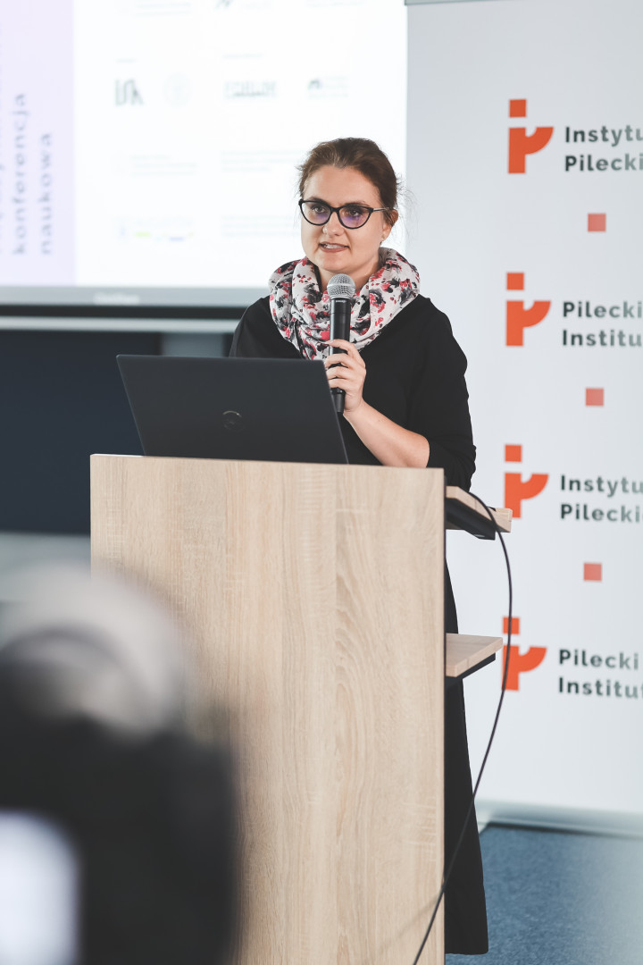 Prelegentka w okularach o ciemnych oprawkach i czarnym stroju, z jasnym szalem w czerwony, kwiecisty wzór, wygłasza referat przy mównicy z laptopem marki Dell. W tle widoczny pionowy rollup Instytutu Pileckiego oraz ekran z logotypami współorganizatorów, partnerów i patronów, w tym wyraźny znak Instytutu Studiów Kobiecych.
