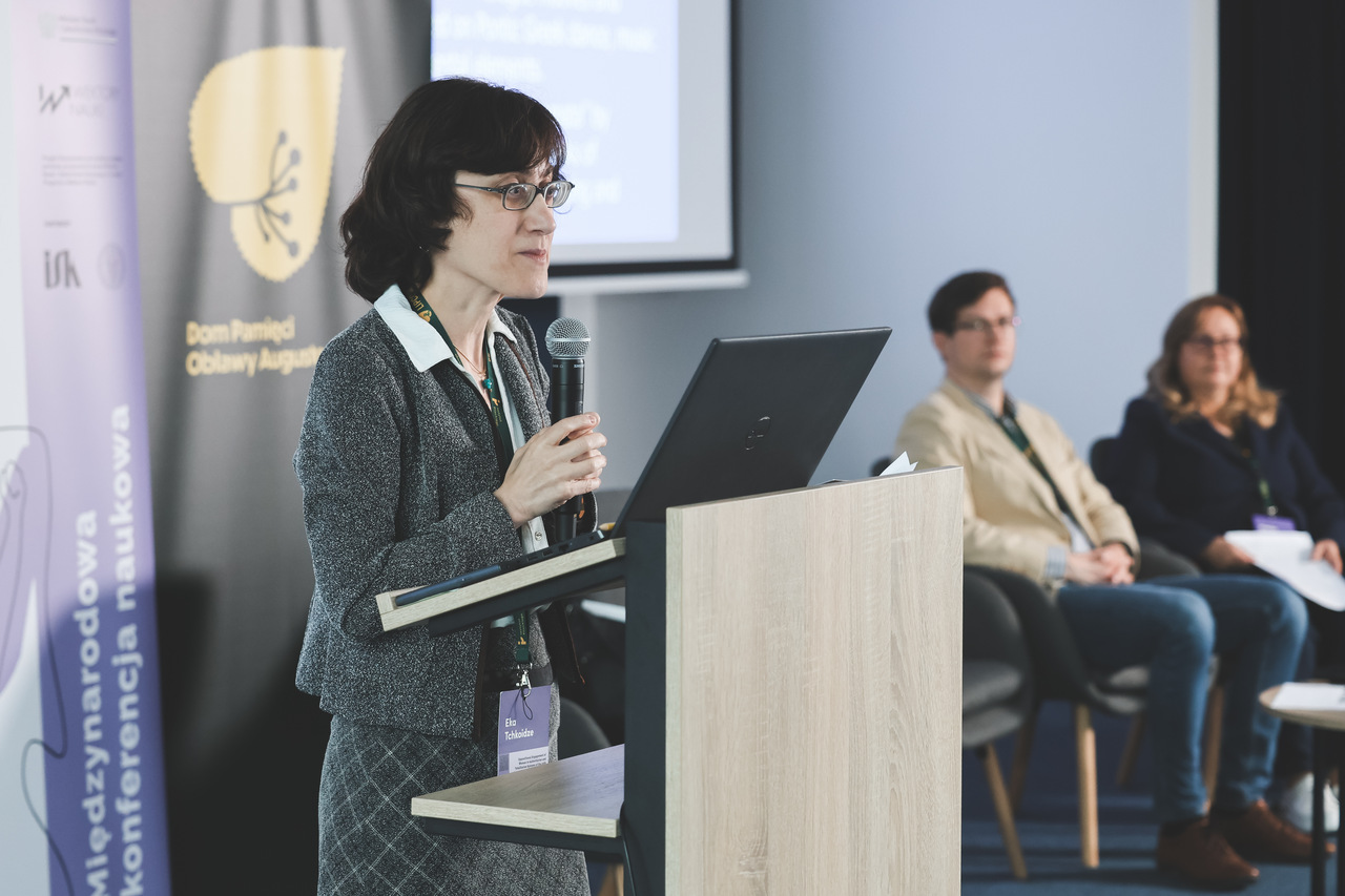 Prelegentka w okularach, szarym żakiecie i kraciastej spódnicy wygłasza referat przy mównicy z laptopem. W tle widoczne rollupy z logotypami programu „Wektory Nauki” oraz Domu Pamięci Obławy Augustowskiej – filii Instytutu Pileckiego. Na drugim planie siedzą pozostali członkowie panelu tematycznego, oczekujący na swoje wystąpienia.